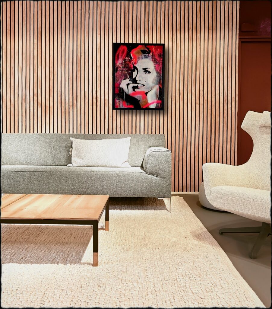 Expressief portretkunstwerk in rood, zwart en wit, gepresenteerd in een zwarte lijst boven een moderne grijze bank. De verticale houten lattenwand, lichte fauteuil en minimalistische inrichting versterken de hedendaagse uitstraling van het kunstwerk. De krachtige kleuren en gelaagde texturen geven het werk een opvallende, artistieke aanwezigheid binnen het interieur.